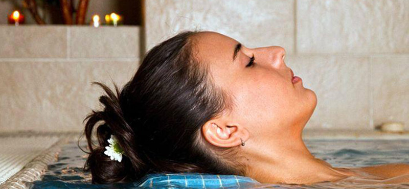 Primer plano del perfil de la cabeza de una mujer con los ojos cerrados recostada en una tumbona dentro de una piscina de hidroterapia.