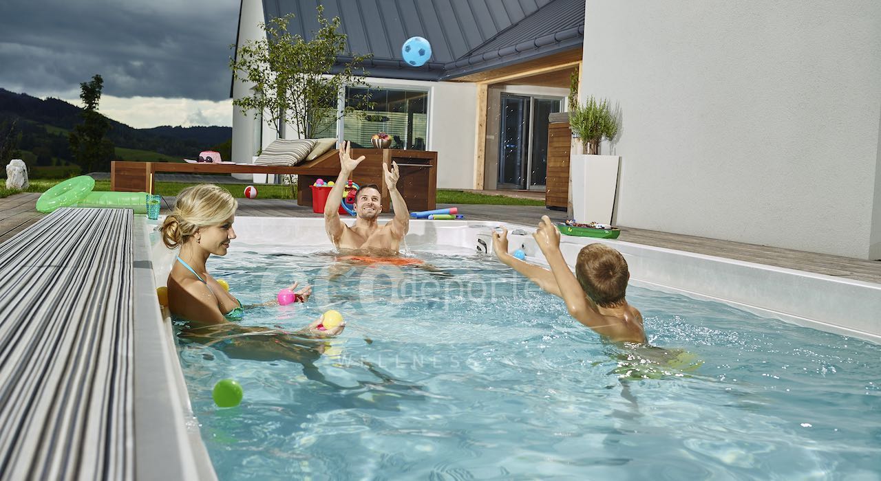 Bañarte en pleno invierno en agua muy caliente ¡El mayor placer! Mujer tomando una sesión de hidroterapia sentada en la escalera de un swim spa exterior colocado entre la nieve de invierno mientras que su compañero, que acaba de llegar de esquiar, la observa